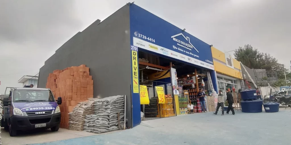 Fachada da loja Multi Soluções Vila Sônia em São Paulo, com estacionamento, displays de materiais de construção, área de logística com caminhões, areia e tijolos.