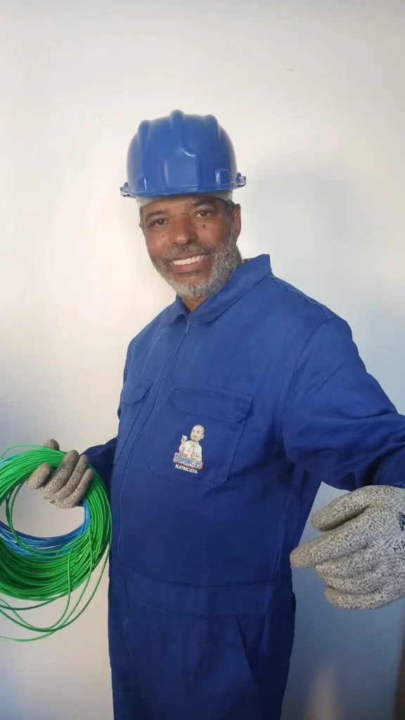 Eletricista confiável em SP segurando rolos de cabo, usando macacão azul personalizado e capacete de segurança, representando o profissionalismo do Seu Domingos Eletricista.